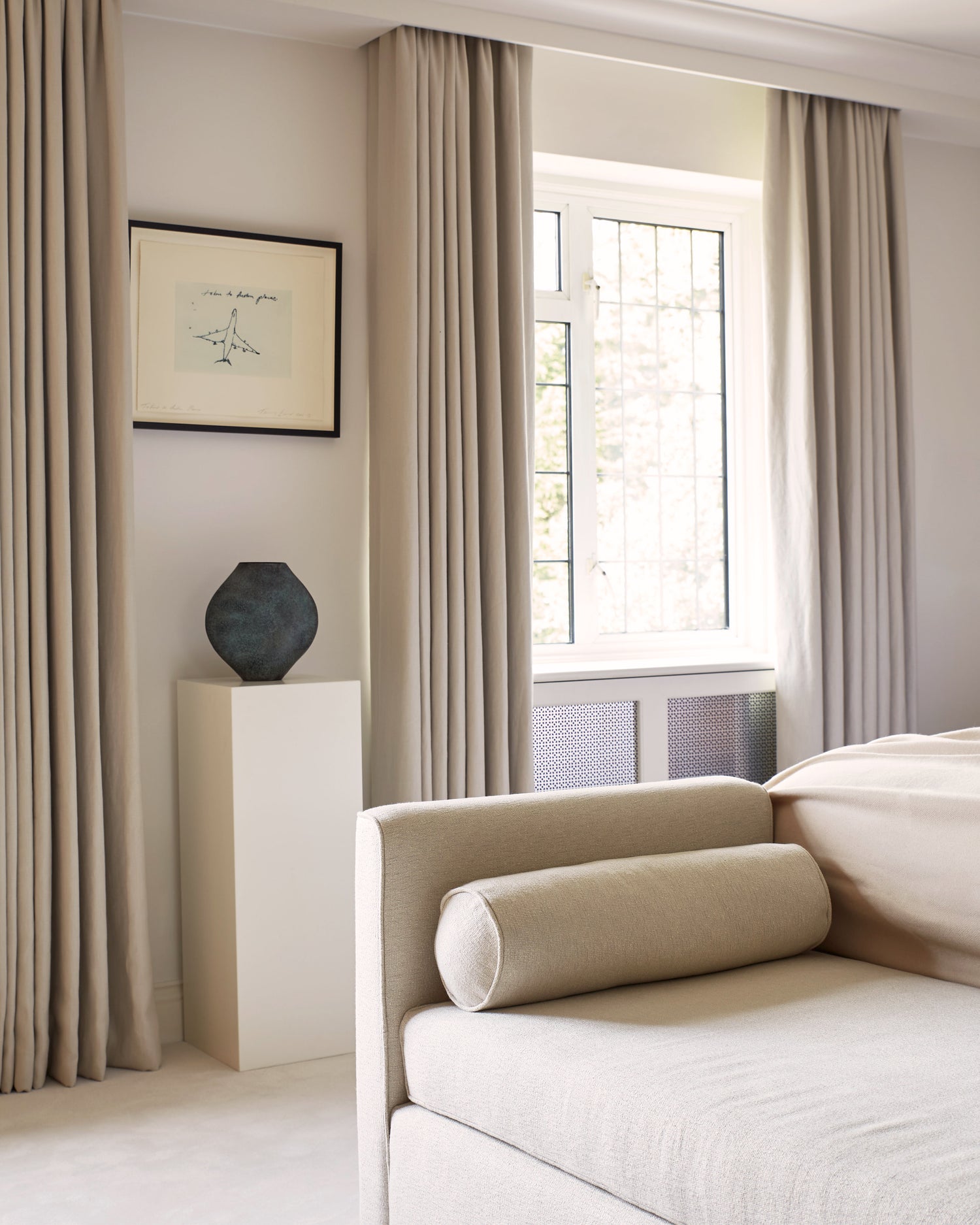 Calm bedroom with cream upholstered chaise, linen curtains, and framed minimal art in London home